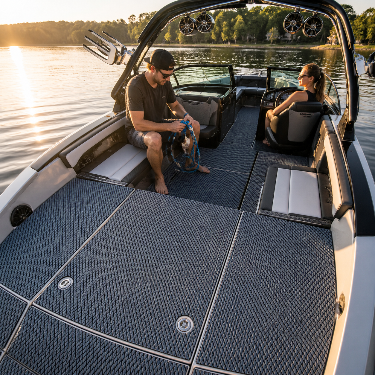 Wake boat cockpit with gray-blue textured flooring, a man coiling rope near the stern, and a woman seated at the helm