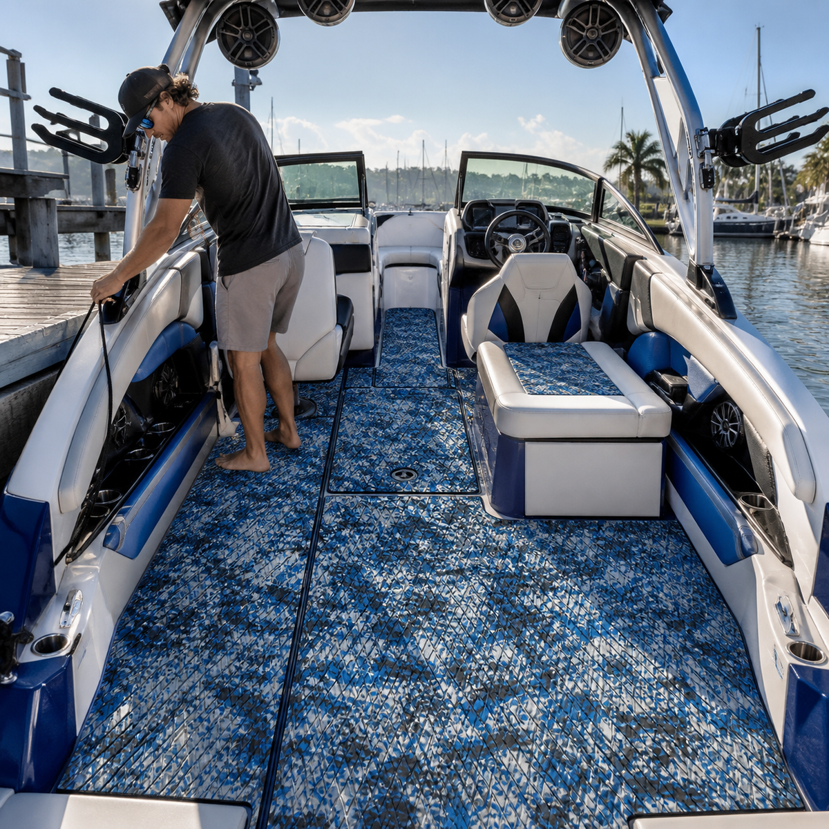 Wake boat cockpit at a marina with blue, white, and black patterned flooring, wraparound seating, a sport tower, and a man standing by the port-side seating