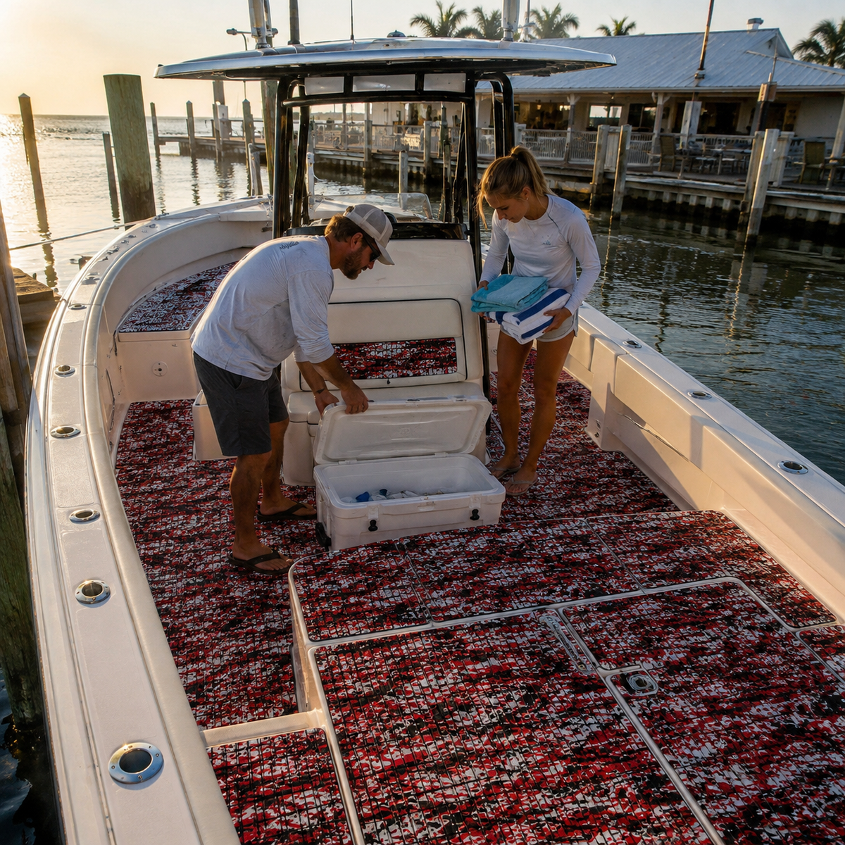 Center-console boat cockpit with red, black, and white grooved flooring, open deck hatches, a cooler, and two adults loading towels at a marina