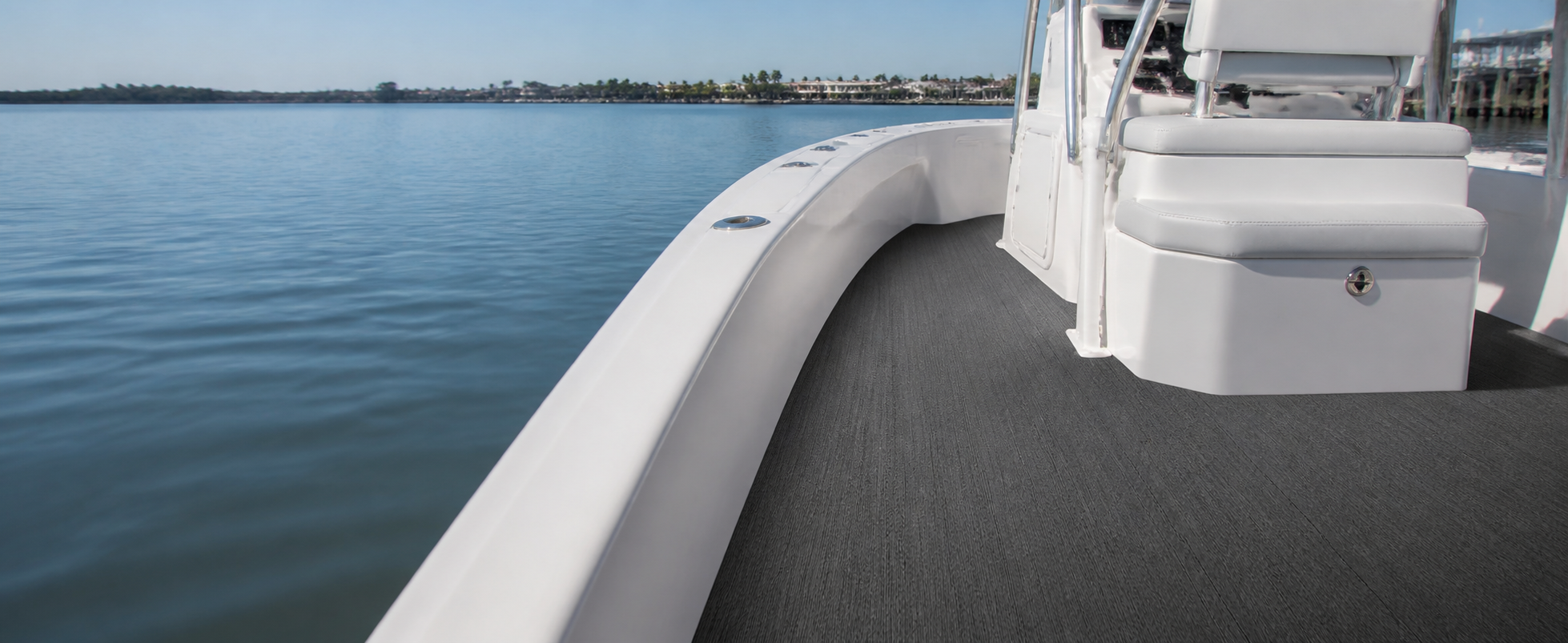 Wide center-console boat deck with gray flooring beside white gunwales and a white console seat, with calm water and a marina in the background