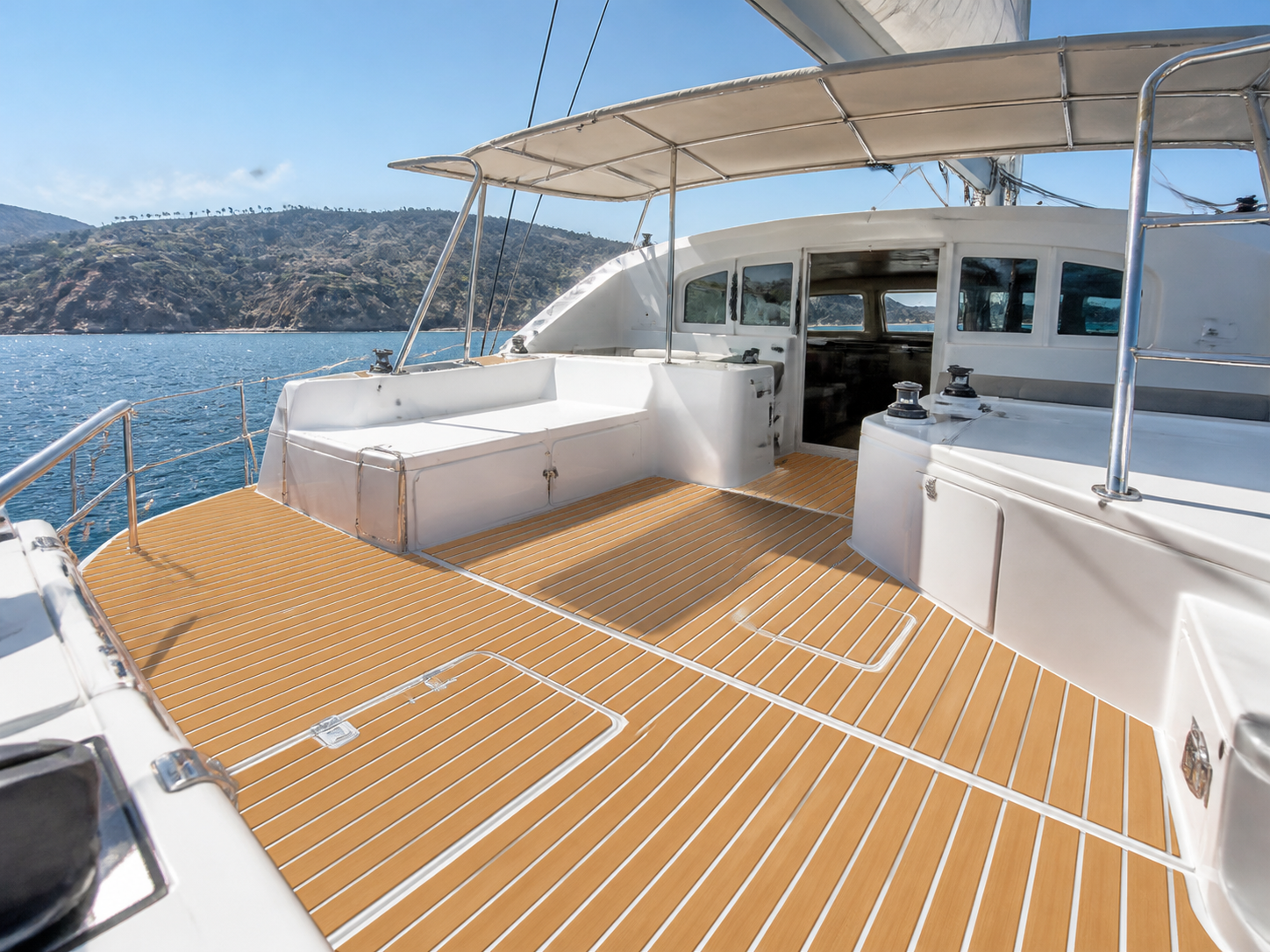 Cruising catamaran cockpit with tan striped deck flooring, white seating, storage hatches, winches, a shaded roof, and blue coastal water